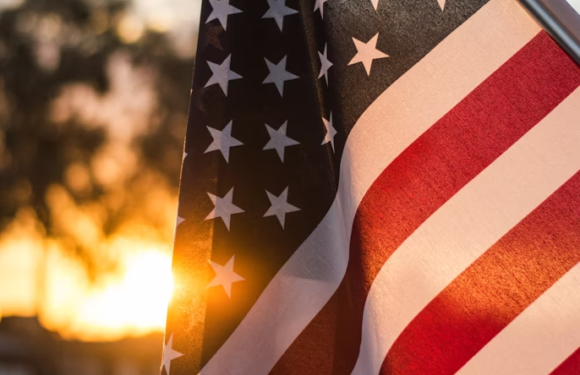 American flag waving at sunset, with a blurred background of trees and sky. The sunlight partially shines through the flag's fabric.
