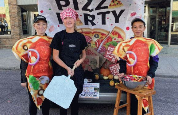 Three children dressed in pizza slice costumes and a girl in a black apron and red checkered hat stand in front of a decorated vehicle with a banner reading "PIZZA PARTY," holding a large cutting board and a bowl of candies.