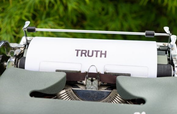 A vintage typewriter with a sheet of paper displaying the word "TRUTH" is set outdoors on green grass.