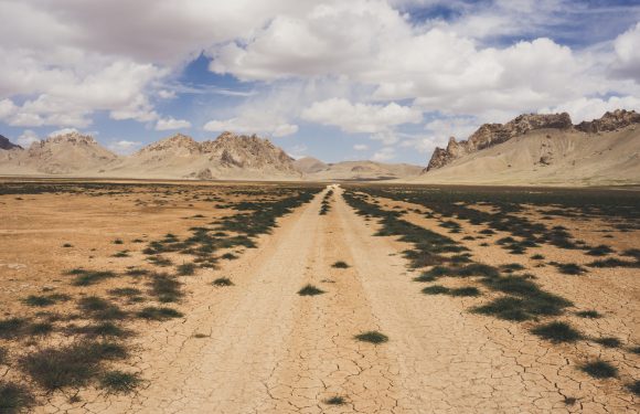 A dry, cracked dirt road extending into the distance across a flat, arid desert landscape with sparse green vegetation and rugged mountains under a partly cloudy sky.