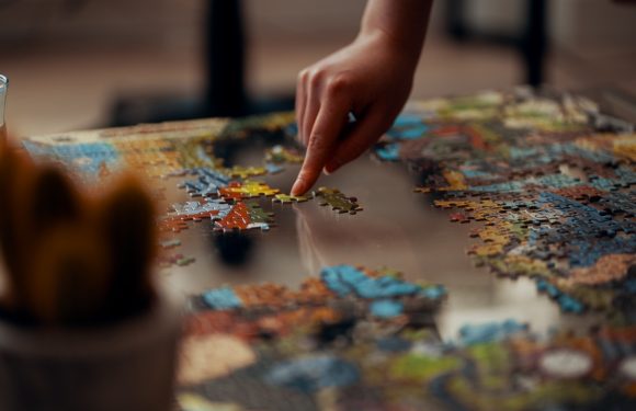 A person's hand is placing a puzzle piece onto a partially completed colorful jigsaw puzzle on a table.