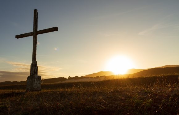 A weathered cross stands on grassy terrain at sunset, with the sun near the horizon behind distant hills, illuminating the sky with warm light.