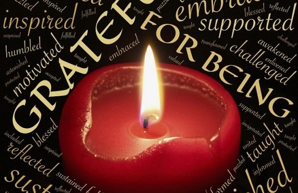 A lit red candle surrounded by words like "gratitude," "supported," "nourished," "connected," and "inspired" in various fonts and orientations, creating a warm, reflective atmosphere.