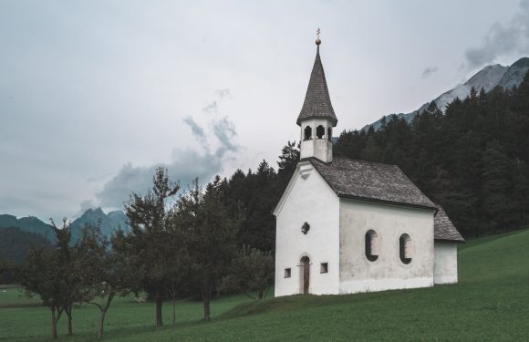 A small white church with a steeple and a cross on top, surrounded by trees and mountains under a cloudy sky.