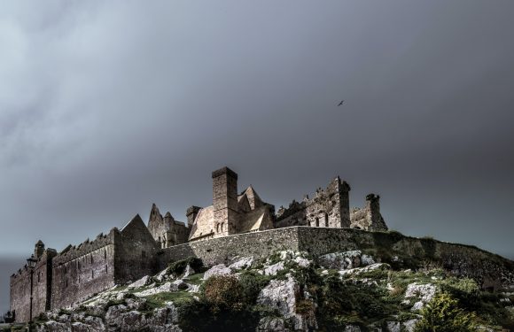 A medieval castle with stone walls and towers sits atop a rocky, grassy hill under a dark, overcast sky. A bird flies in the distance.
