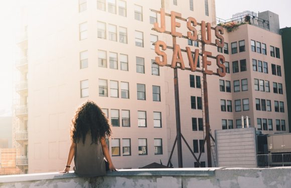 A woman with curly hair sitting on a rooftop ledge, looking at a large "JESUS SAVES" sign on a building in an urban setting during daylight.