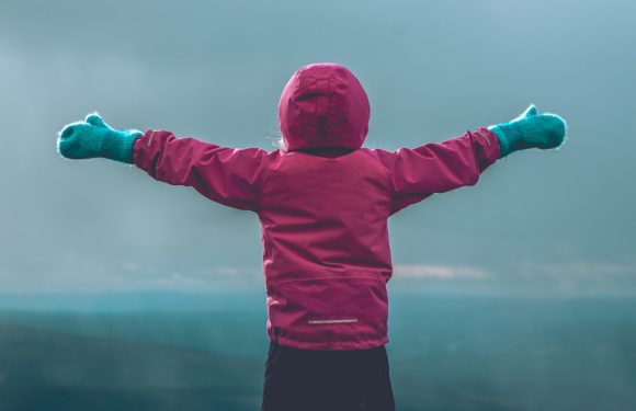 A child in a pink jacket and blue gloves stands outdoors with arms outstretched, facing away, on a cloudy day.