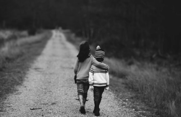 A woman and a child walk together down a dirt road surrounded by grass and trees, with the woman gently holding the child's shoulder.
