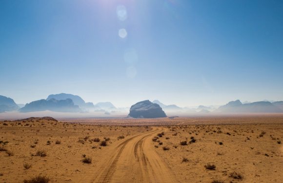 The image shows a desert landscape with a dirt track leading toward large rock formations in the distance, under a clear blue sky with the sun high overhead.