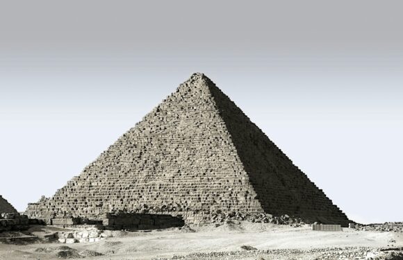 A large stone pyramid sits in a desert landscape, with a clear sky above and scattered stones and ruins at its base.