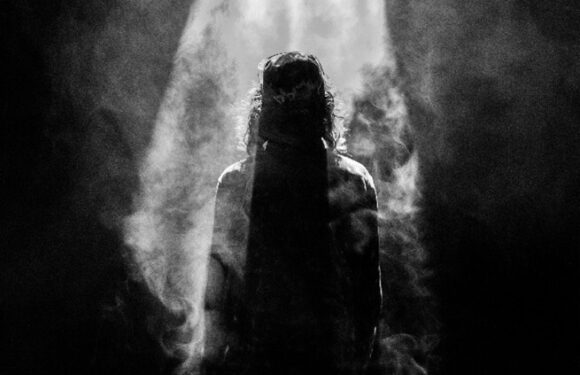 A person with long hair stands in a spotlight surrounded by smoke, creating a mysterious, dramatic atmosphere in a dark setting.