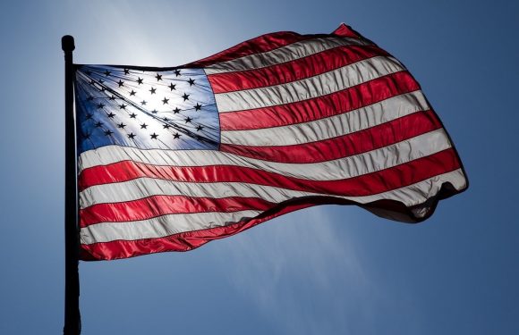 American flag waving in the wind against a blue sky with sunlight behind it.