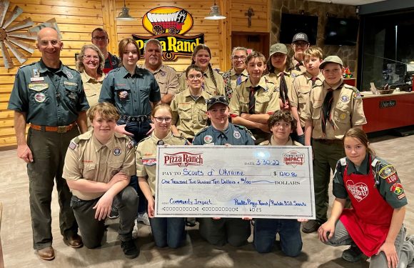 Group of scouts and leaders holding a large check made out to Scouts of Ukraine for $1,210.80 at a Pizza Ranch restaurant, with wood-paneled walls and a neon sign in the background.