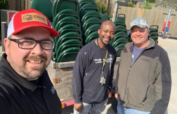 Three men outdoors, smiling, standing in front of stacked green chairs on a truck. One wears a red and white cap with a logo, the other two wear casual jackets.