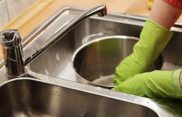 Hands wearing green rubber gloves are washing a round metal pizza pan in a stainless steel kitchen sink.