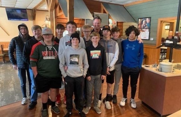 Group of boys and an adult man, some wearing casual clothes and one in a Pizza Ranch uniform, standing indoors on a wooden floor, in a restaurant setting.