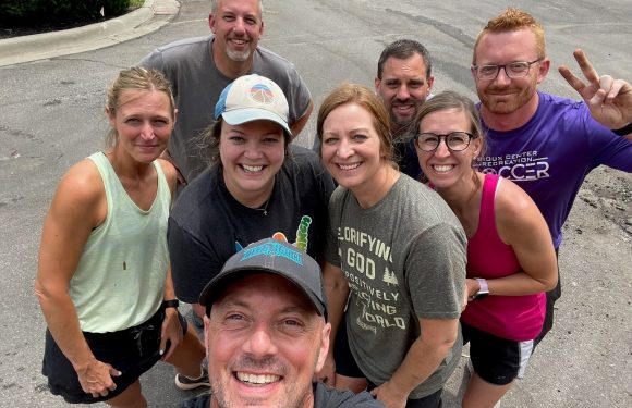 A group of eight people taking a selfie outdoors, smiling, some wearing athletic clothing and glasses, with a man in the front holding the camera.