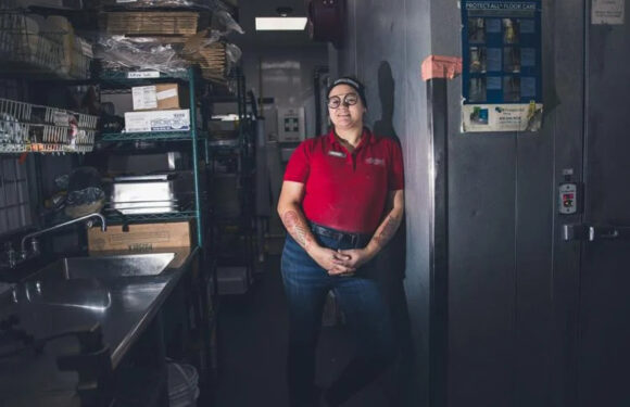 A woman wearing glasses and a red Pizza Ranch uniform stands with her hands clasped in a kitchen with stainless steel shelves, a sink, and storage areas.