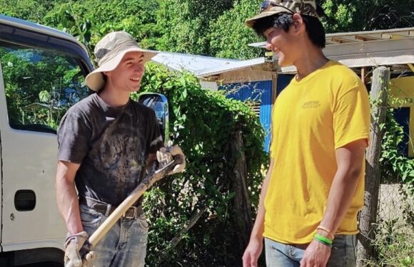Two young people outdoors, smiling and talking, one holding a shovel, with lush green trees and a small building in the background.