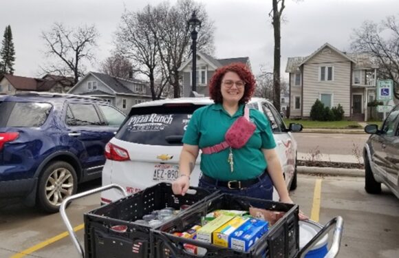 A woman with curly red hair, glasses, and a green shirt stands in a parking lot with a cart filled with groceries, smiling at the camera. She has a pink crossbody bag.