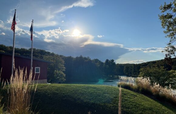 A serene lakeside scene with a grassy hill, trees, two flags, a small building, and the sun peeking through cloud cover over the water.