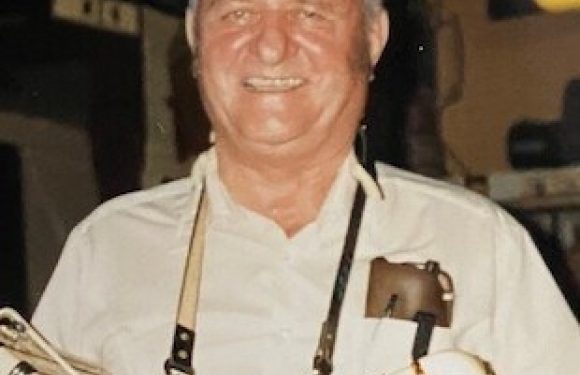 Smiling man in a white shirt with a pen pocket, wearing a neck strap, standing behind a display of cookbooks or similar items, in an indoor setting.