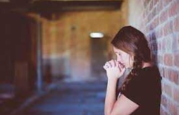 A woman with brown hair and glasses looks down thoughtfully, standing near a brick wall in a dimly lit corridor.