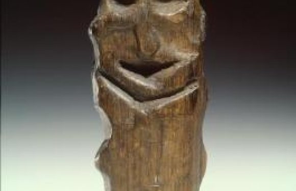 Wooden carved figure with a smiling face, rounded eyes, and a simple mouth, set against a dark background with a light gradient at the bottom.