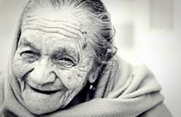 A smiling elderly woman with gray hair and wrinkles, wrapped in a cozy blanket, sitting indoors with a warm, cheerful expression.