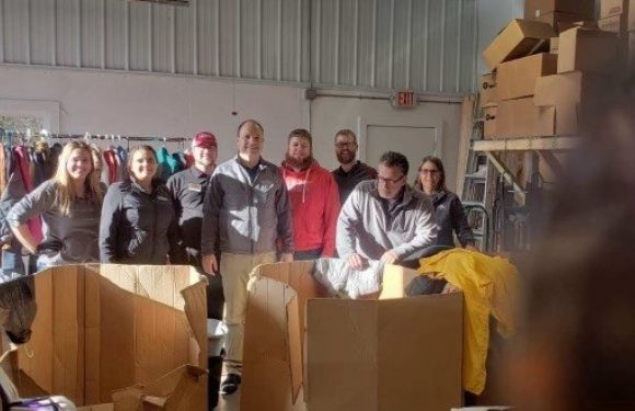 Group of eight people standing inside a warehouse among shelves and cardboard boxes, some wearing matching work uniforms, with a clothing rack on the left.