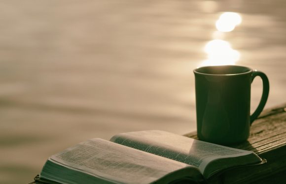An open book and a mug are placed on a wooden dock by a body of water at sunset, with reflections of sunlight shimmering on the water's surface.