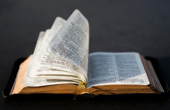 An open Bible with pages slightly curled, resting on a dark surface with a plain background.