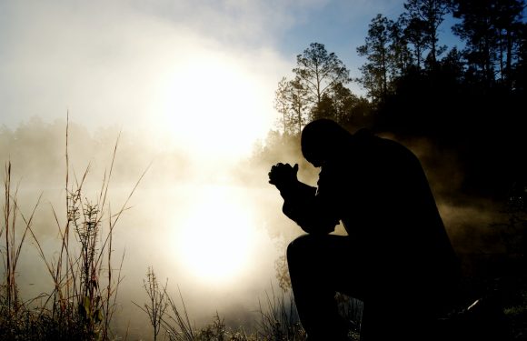 A person in a uniform kneels by a foggy riverbank at sunrise, hands clasped in a prayer or reflection, with trees in the background.