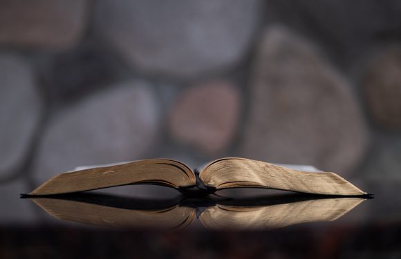 An open, well-worn book with slightly curled pages, resting on a reflective dark surface, against a background of rounded stones.