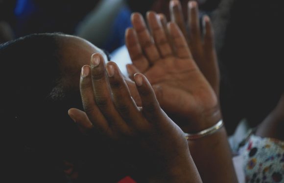 A person with dark skin and a bangle on their wrist holds their hands up with palms facing outward, possibly in prayer or a moment of reflection.
