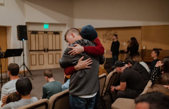 Two men embrace in a hug in a room with seated audience members. One wears a beanie and a red hoodie, the other a gray jacket, with a wooden wall and door in the background.