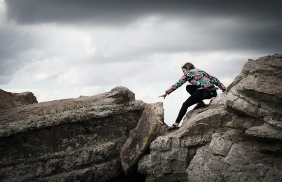 A woman in a floral jacket is climbing over large rocks on a cloudy day, reaching out with her right hand to steady herself.