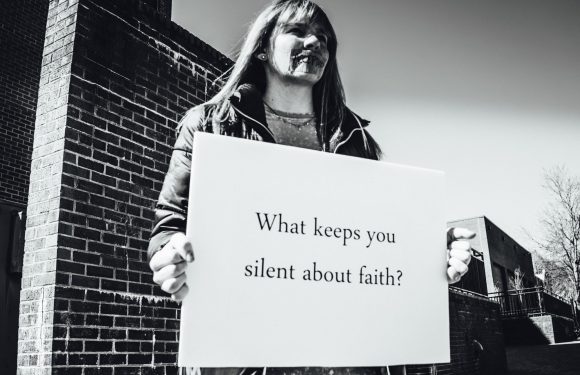 A woman holding a sign that reads "What keeps you silent about faith?" standing outdoors near a brick building with a clear sky in the background.