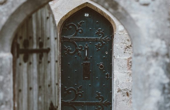 A weathered stone archway with a wooden gate leads to a black metal door with ornate scrollwork and the number 22, framed by aged stone walls.