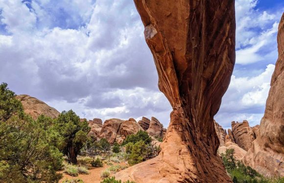 A tall, narrow rock formation in a desert landscape with scattered green bushes and trees, under a partly cloudy sky.
