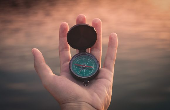 A person's hand holding a compass with an open lid, with water and a sunset in the background. The compass shows north, south, east, and west directions.
