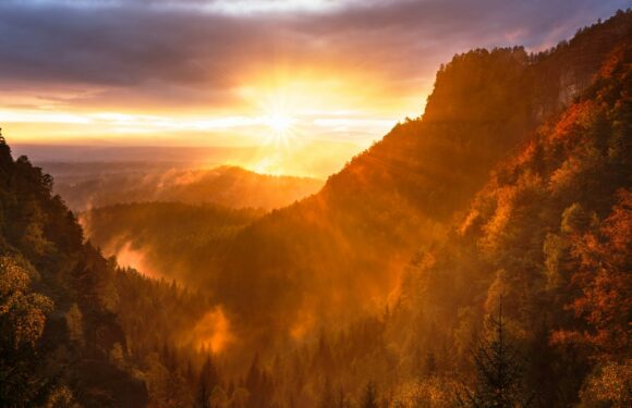 A scenic mountain landscape during sunset with the sun's rays shining through the sky, illuminating forested valleys with autumn-colored trees and mist.