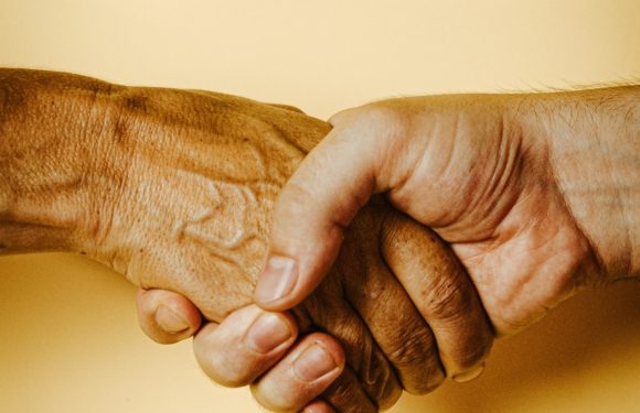 A close-up of two hands shaking, one with darker skin and the other with lighter skin, against a plain yellow background. The person on the right is wearing a Pizza Ranch uniform.