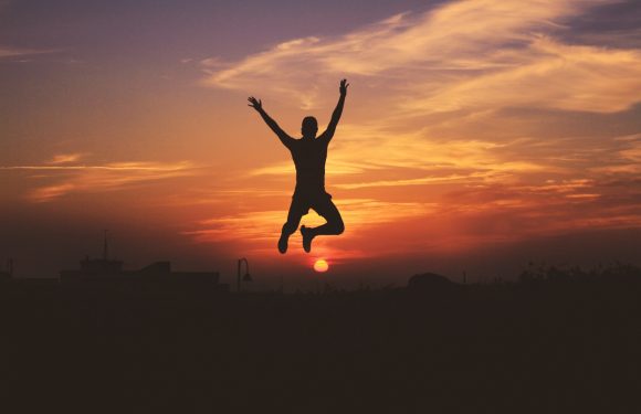 A person jumping in the air with arms raised against a colorful sunset sky, silhouette with urban elements and streetlights faintly visible below.