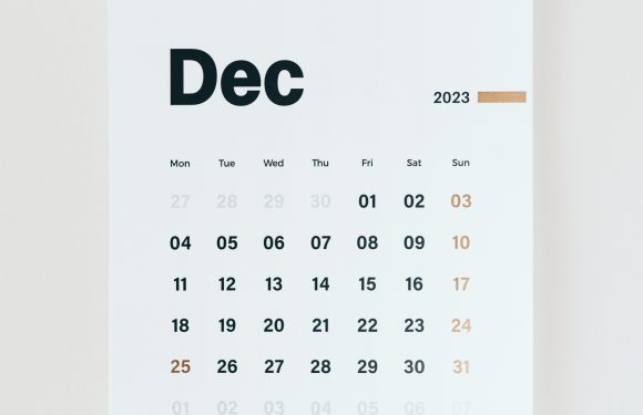 A wall calendar for December 2023 with black and brown text, white background, and a black and beige clip at the top, hung on a plain wall.