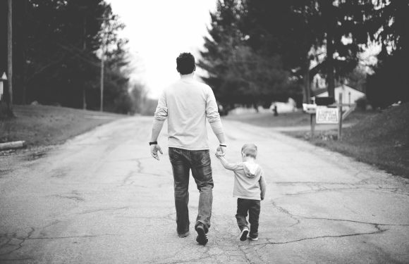 A man and a boy walk hand in hand down a cracked, empty street lined with trees and houses. The image is in black and white.