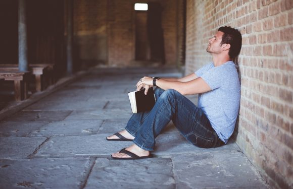 A man sits on the floor against a brick wall, holding a closed book, wearing a T-shirt, jeans, flip-flops, and a watch, appearing contemplative.