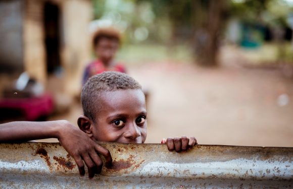 A young boy with short curly hair peeks over a rusty metal barrier, with a blurred girl in a hat and colorful outfit in the background outdoors.