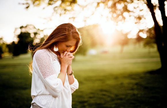 A woman with long hair, wearing a white crochet top and earrings, stands outdoors with her hands clasped in prayer, illuminated by warm sunlight in a park.
