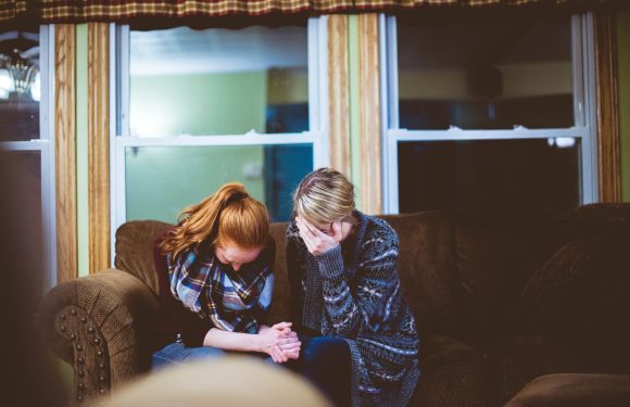 Two women sitting on a brown couch, appearing upset, with one covering her face and the other holding her hands, in a room with green walls and windows.
